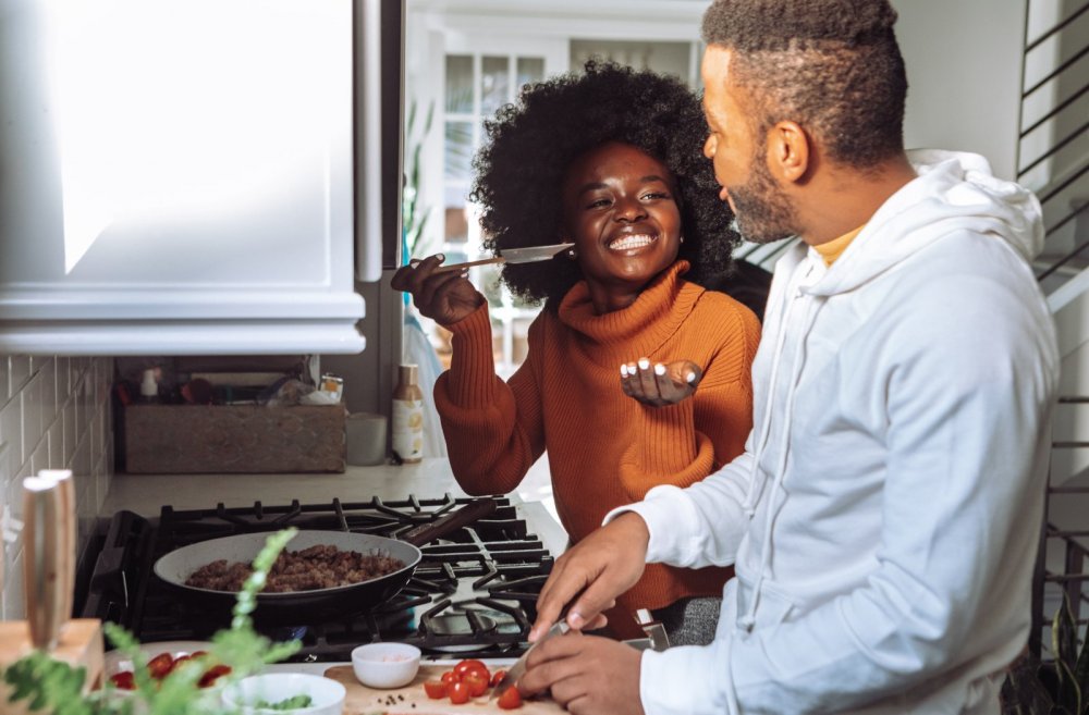 Selbstfürsorge Tipps: Probier mal was Neues - Bild zeigt ein junges Paar lachend beim Kochen am Herd. 